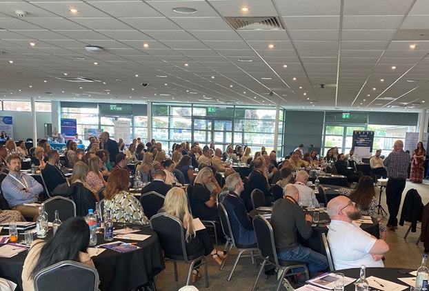Wide shot of a room full of delegates in a conference suite whilst a member of the team stands at the front addressing the room.