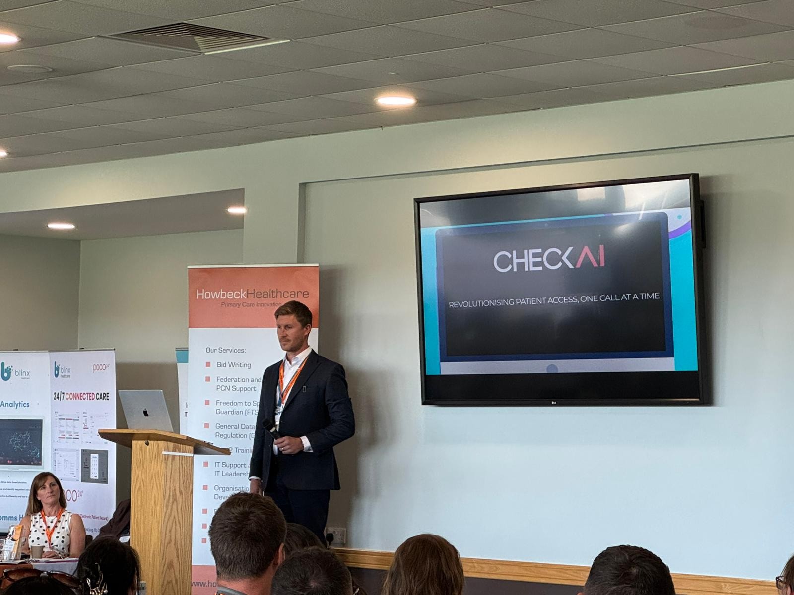 A male speaker dressed in a dark suit from CheckAI stood on stage during a conference addressing the audience.
