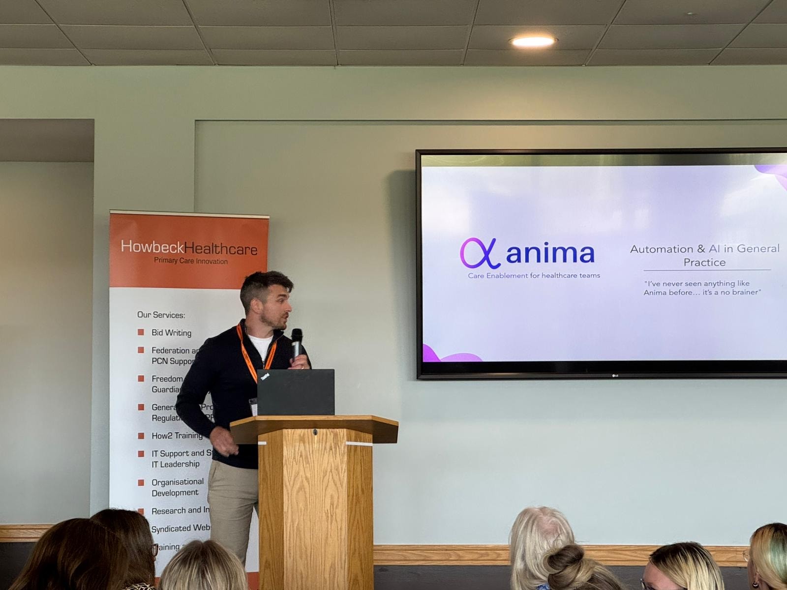 A male speaker dressed in stone coloured trousers and a dark jumper from Anima stood on stage during a conference addressing the audience.