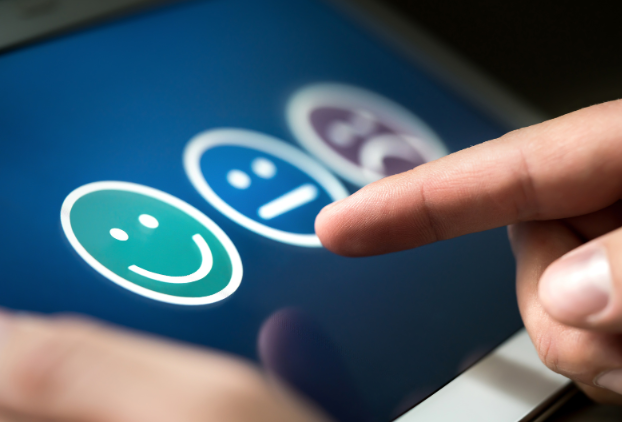 A finger pointing at a blue screen with three emoji faces, one with a happy face, one with a neutral face and one with a sad face.